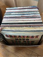 Top view of a box containing approximately 30 assorted vintage vinyl record albums. The album covers show various artists and titles, including Greatest Hits of the New Country and Broadway Hits, along with other classic rock and country artists.