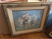 Painting of a floral arrangement with daisies in an ornate beige frame with floral corner details.