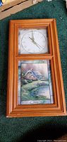 Front view of the wooden wall clock showing clock face with Roman numerals and scenic landscape picture below.