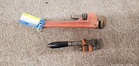Photo showing two pipe wrenches: a large red multi-purpose pipe wrench and a smaller pipe wrench with a black handle and orange jaw, placed on a carpeted floor.