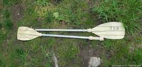 Two-piece kayak paddle lying on grass, showing beige plastic paddle blades connected by aluminum shafts, blades marked with JSB and 95C