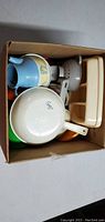 Overhead view of a box filled with assorted plastic and ceramic pretend play kitchen food items and utensils.