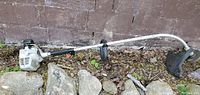 Side view of grey ECHO gas powered weed trimmer wacker showing full length with shaft, motor, handle, and cutting head on the ground with some rocks and leaves around.