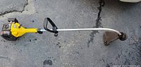 Side angle image of yellow and black MAC 2816 gas powered weed trimmer with curved shaft and protective blade guard showing wear and rust on asphalt.