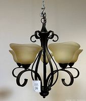 Front view of chandelier showing black metal scroll frame and four beige frosted glass floral-shaped lampshades.