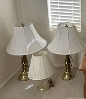 Two brass table lamps and one crystal desk lamp grouped together on carpeted floor near window with blinds.