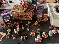 Photo showing a woven basket labeled 'JOY', surrounded by various Christmas figurines including reindeer, snowmen, elves, toy soldiers, and miniature Christmas trees, as well as boxed ornaments in purple packaging.