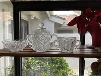Crystal creamer set with three pieces placed on a wooden shelf by a window. Larger canister has a cracked lid.