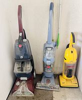 Four vacuum and floor cleaning machines lined up against a wall, showing maroon, blue, yellow, and gray units.