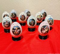 Front view showing the dozen Hummel collectible eggs with various Hummel child artwork, each on black stands against a red cloth background.