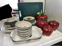 Full view of stacked cream-colored dishes with dark green rims and apple motifs, two rectangular platters, and three apple-shaped canisters with green leaf lids.
