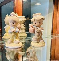 Photo of two Precious Moments porcelain figurines, boy with bag over shoulder and girl with fishing rod, displayed in a glass cabinet.