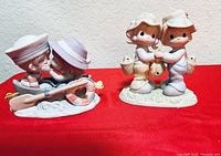 Photo showing two Precious Moments figurines on red cloth background: girl kissing boy on boat with guitar, and fishing boy and girl with basket and fish.