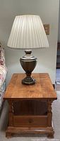 Single brass table lamp with beige pleated shade on wooden end table near sofa.