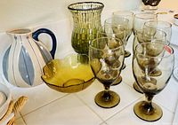 Photo showing six brown water goblets, green textured glass vase, olive colored glass bowl, and blue-white striped ceramic pitcher with handle on a white surface.