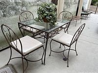 Wrought iron patio set showing four chairs with cushioned seats around a rectangular glass top table, with a faux potted plant in the center, all on outdoor concrete floor