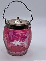 Full view of cranberry glass jar with metal lid and handle, showing the overall shape, color, and handle.