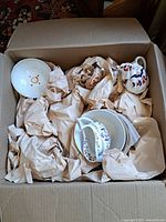 Box containing several antique china pieces including tea pots, plates, and cups wrapped in brown paper.