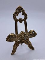Front view of small vintage brass display stand showing detailed ornate cutout floral and scrollwork design with raised platform from front.