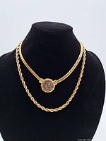 Front view of two gold-tone necklaces displayed on a black bust; one is a twisted rope chain, the other has a flat chain with a round vintage coin pendant.