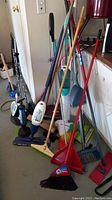 Photo showing multiple brooms with wooden and plastic handles, dustpans, and part of the Bona floor cleaner system clustering near a door and cabinet.