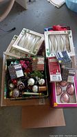 Photo showing several boxes including boxed white glass ornaments labeled 'Alderbrook', a box with mixed holiday decorations, unopened new LED light sets, and a box with pink glass ornaments.