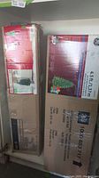 Two boxed artificial Christmas trees stacked horizontally, boxes showing brand names Holidaytime and General Electric, with tree dimensions and images on packaging.