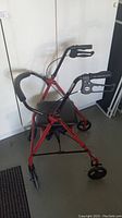 Full view of red folding walker with black padded seat, four wheels, upright handles with hand brakes, and under seat storage compartment.