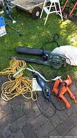 Overview of all garden tools including blower/vac, reciprocating saw with manual, hedge trimmer, power rake, and power cord placed on ground outdoors.