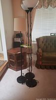 Three floor lamps positioned in a corner near a wooden cabinet and green upholstered chair. Two lamps have white glass torchiere shades, one has a beige fabric shade.