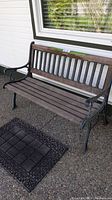 Front angle showing wood slatted bench seat and back framed by ornate iron armrests and legs. Floor mat placed beneath front of the bench.