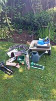 Photo showing assorted garden tools including watering can, sprinklers, fittings, lightweight plastic wheelbarrow with rust on steel frame, and hoses.
