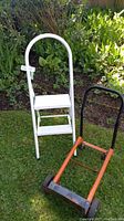 White folding two-step metal ladder with curved handle and red and black hand cart/hand truck on grass