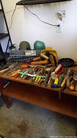 Wide view showing a variety of garden tools laid out on a wooden table. Includes pruners, sheers, gloves, and other hand tools with wooden and plastic handles.