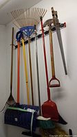 Photo of various yard tools including blue plastic snow shovel, orange and blue leaf rakes, garden broom with green handle, garden edger with wooden handle hung on wall