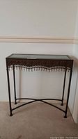 Ornamental table with rectangular glass top and dark metal frame, shown standing against a wall.