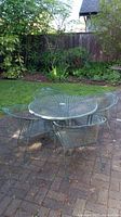 Green wrought iron round patio table with mesh top and decorative floral edge, with four matching chairs on brick patio.