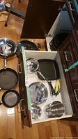 Overview of pots, pans and roasters in an open drawer and on the floor including a square grill pan, wok pan, round roasters, frying pans, and smaller cookware