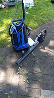 Full view of Simoniz pressure washer with attached hose and power cord, and Jobmate leaf blower on ground outdoors.
