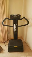 Front view of the Progression Fitness vibration machine showing its black standing platform, vertical column with brand name, and curved handle grips.