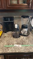 Lot of three small kitchen appliances on countertop: Mr. Coffee Tea Cafe coffee maker, Ninja food processor, Oster blender.