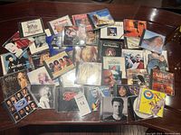 Photo showing a wide assortment of music CDs on a wooden table surface, including pop music artists, musicals, and various genres.