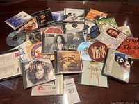 Photo showing a collection of approximately 21 assorted music CDs and albums of mixed artists and genres, arranged on a flat surface.