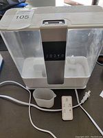 White humidifier with clear water reservoirs and control panel, power cord and remote control on table next to it, also small white measuring cup visible at humidifier base