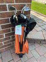 Kids golf set showing orange bag with multiple golf clubs leaning against brick wall on outdoor paving.