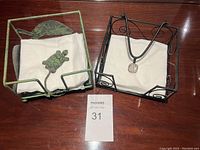 Two metal napkin holders on a wooden surface, one green with a turtle design and one black with decorative swirls and a clear knob, both holding white napkins.