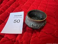 Front angled view of round bronze incense burner showing detailed floral relief and patina on red fabric background.