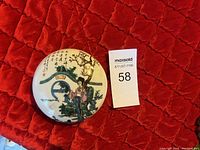 Top view of round porcelain ink pad box lid showing Chinese figures, calligraphy and floral motifs.