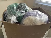 Cardboard box filled with multiple plastic bags containing assorted yarn skeins in varied colors including green, black, and purple.
