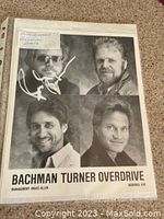Close-up showing Randy Bachman's autograph in white ink on the top left image of the photo. Handwritten note taped to sleeve stating 'Autographed Bachman Photo'.